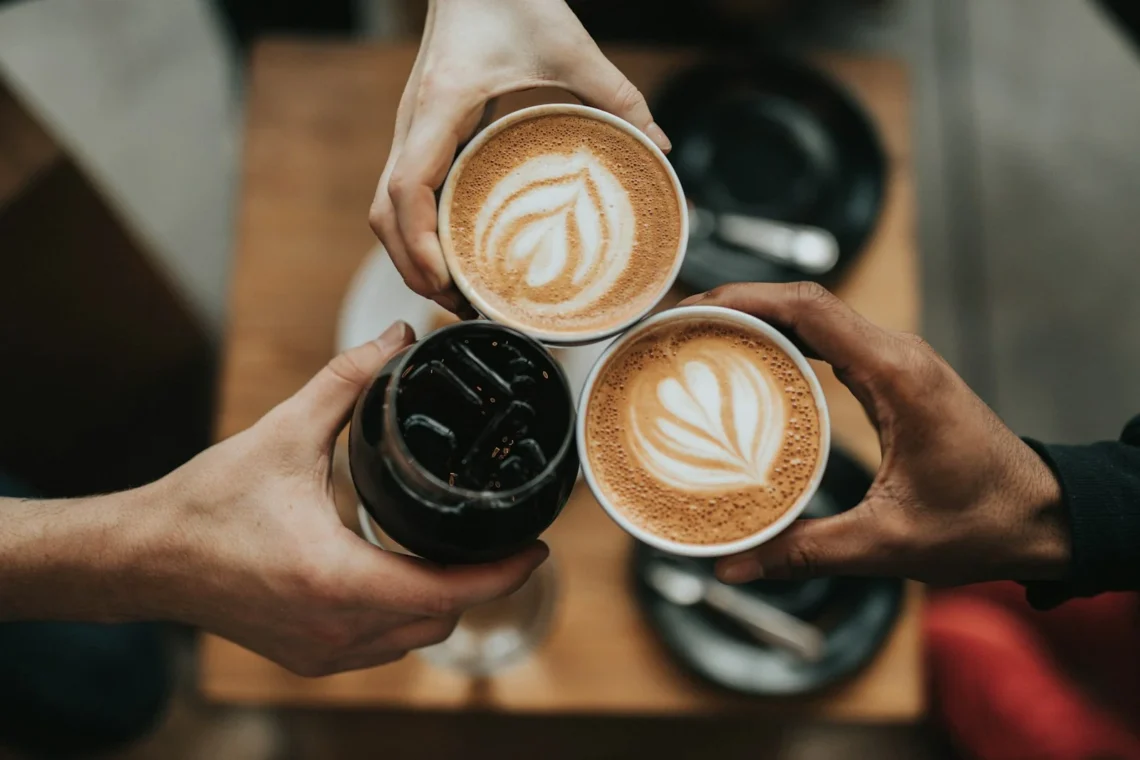 Tre persone che uniscono tazze di caffè e un bicchiere di iced coffee in un brindisi sulla tavola
